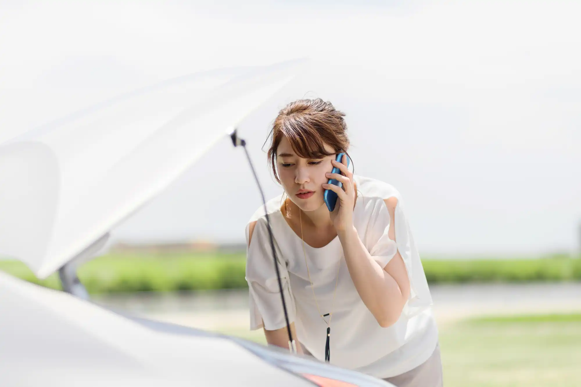 車のボンネットを開けて覗き込みながら電話する女性